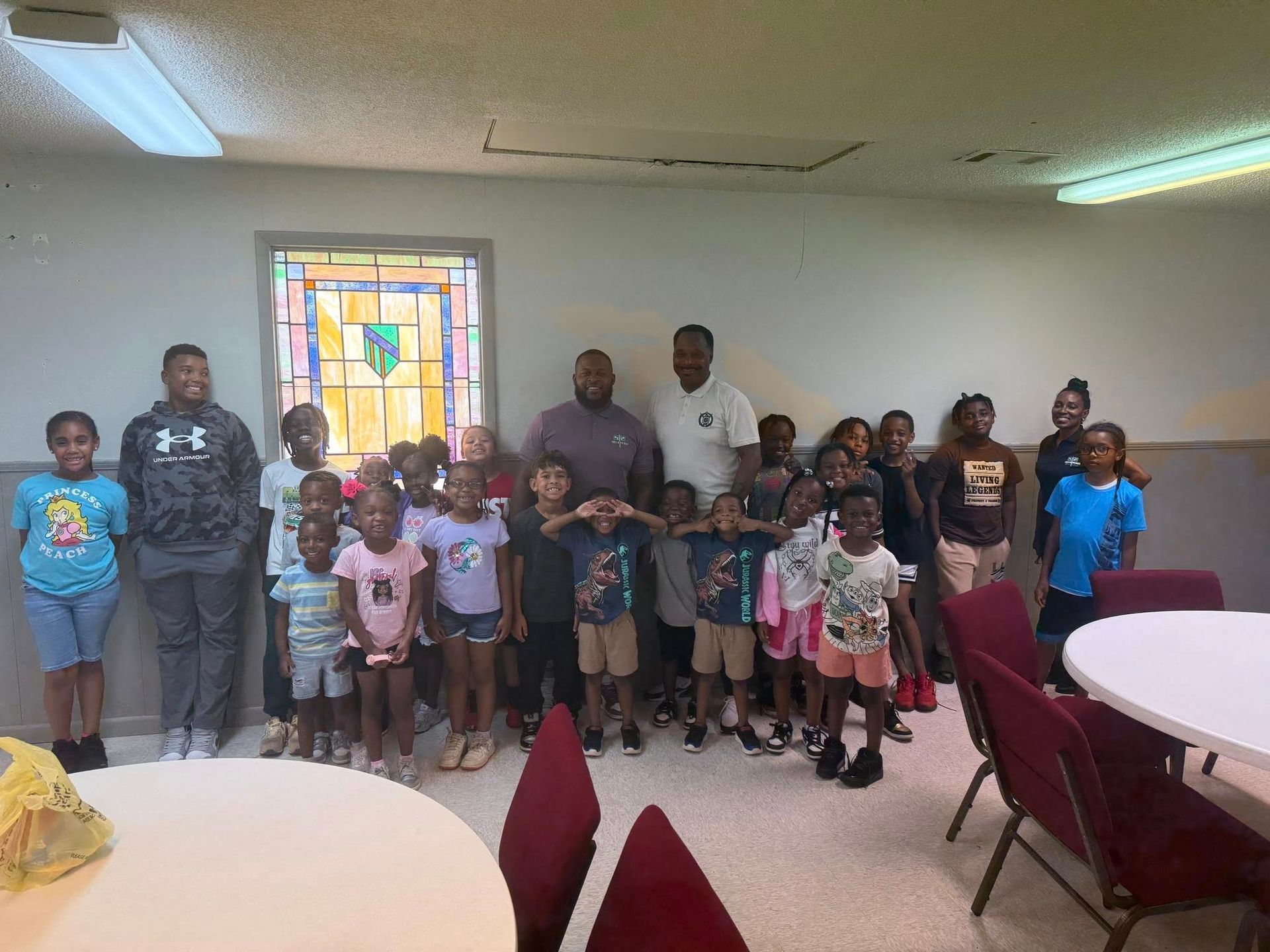 Group of children and adults posing indoors in front of a window. Some people cover eyes.