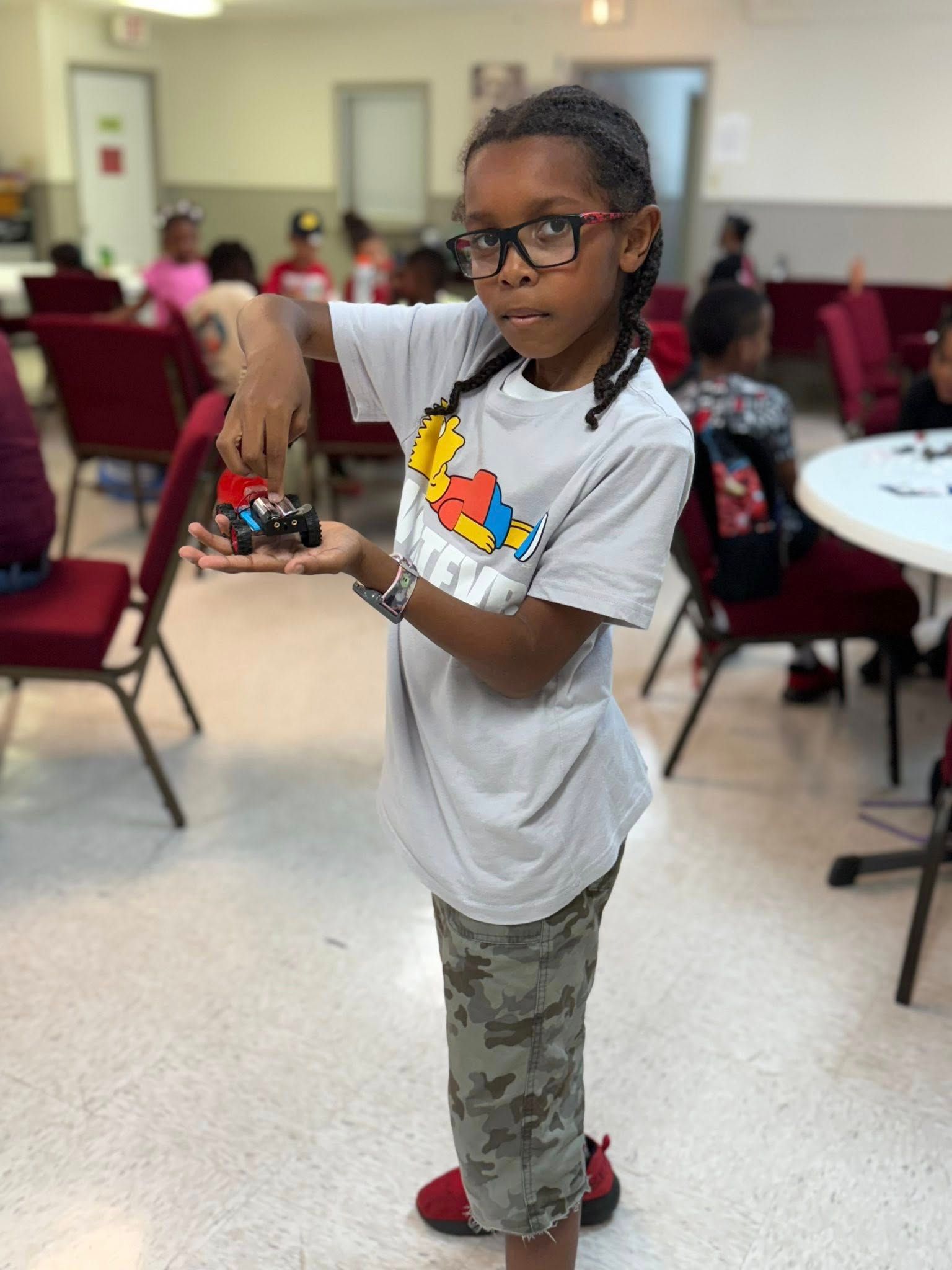 Young person holding up a toy car. Indoor setting, wearing glasses and casual clothing.