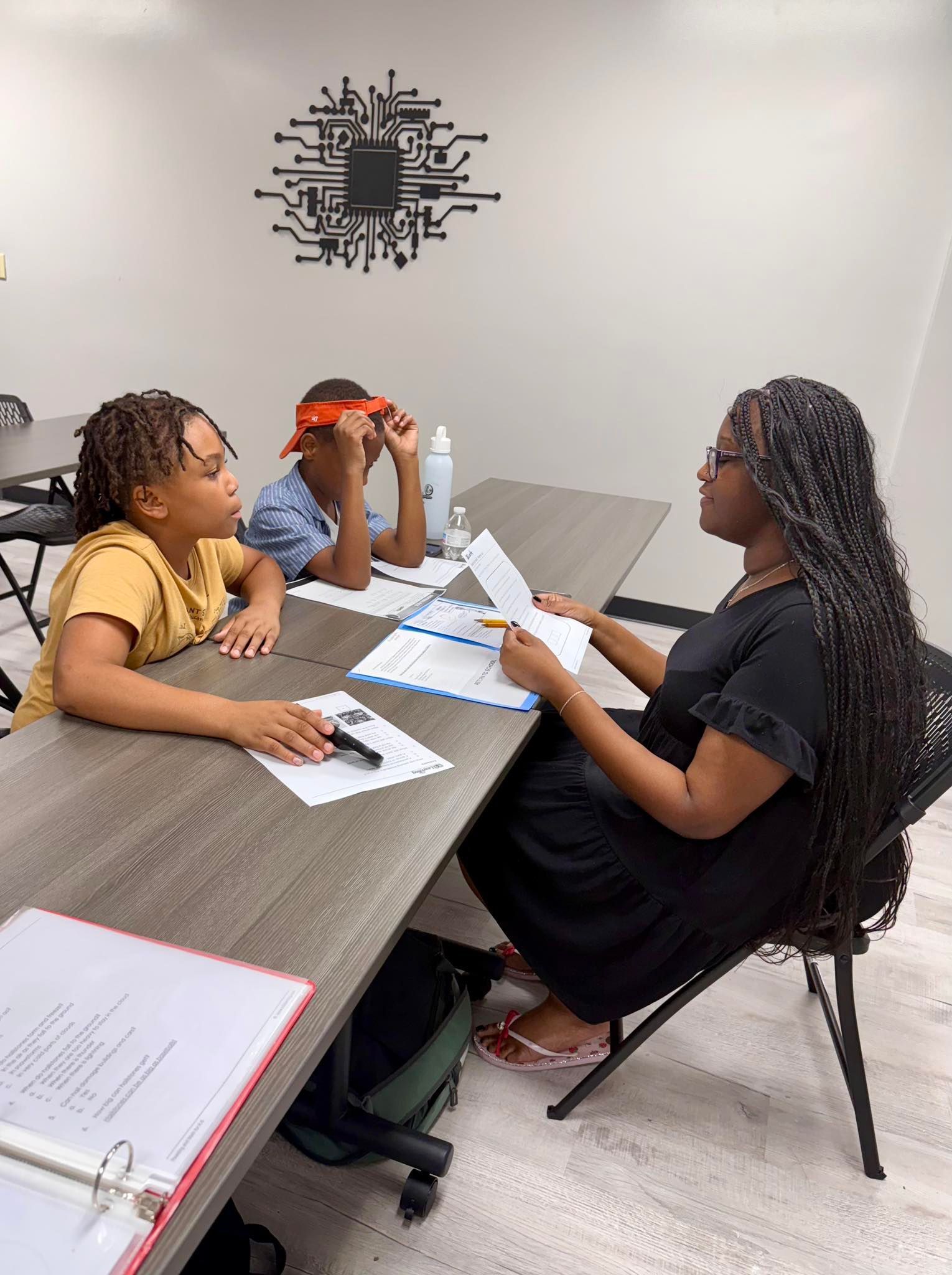 Three people sit at a table in a classroom. A person with braids holds papers, speaking to two students.