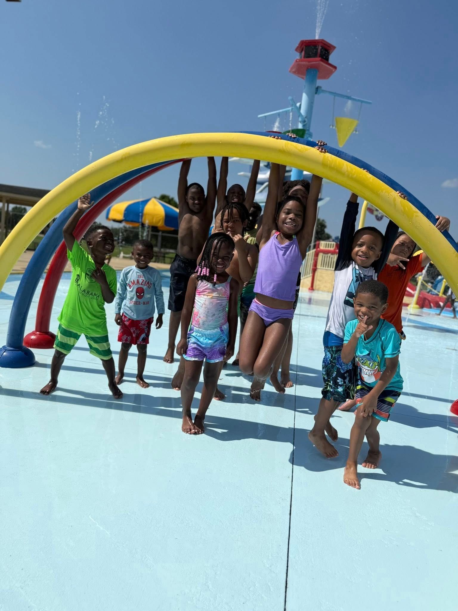 Children playing in a splash pad on a sunny day. They hang from yellow arch, smiling and splashing.