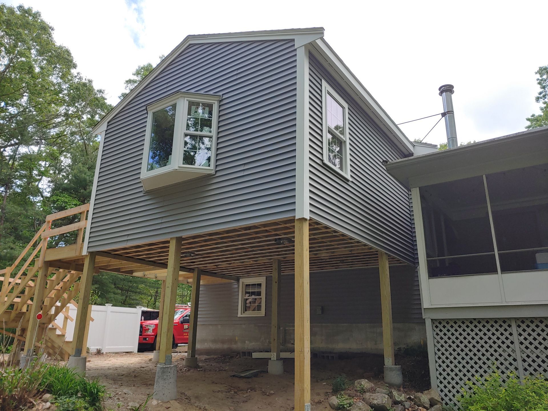 A gray, elevated house structure on wooden stilts with a bay window and an attached wooden staircase in a wooded area.