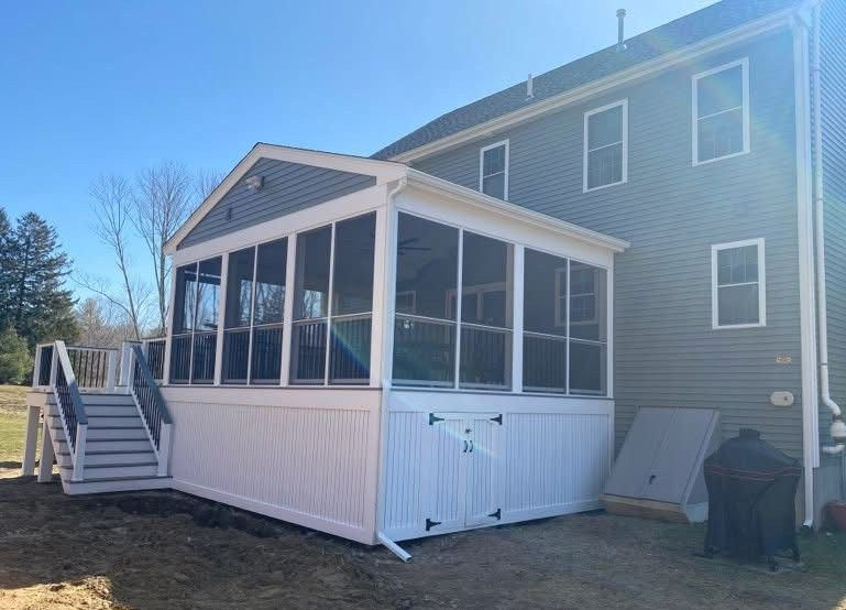 A grey house with a white-framed screened-in porch, wooden steps leading to the yard, and a white storage door underneath.