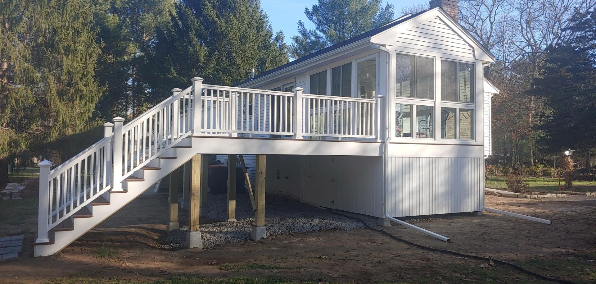 A white sunroom addition featuring a deck with white railings and stairs, built on wooden posts over a graveled area.
