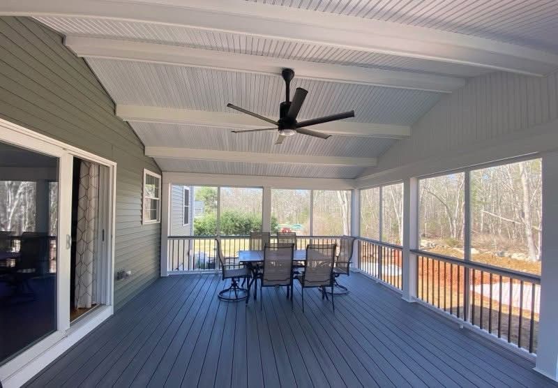 A covered outdoor porch with dark gray decking, screened walls, a ceiling fan, and a patio dining set.