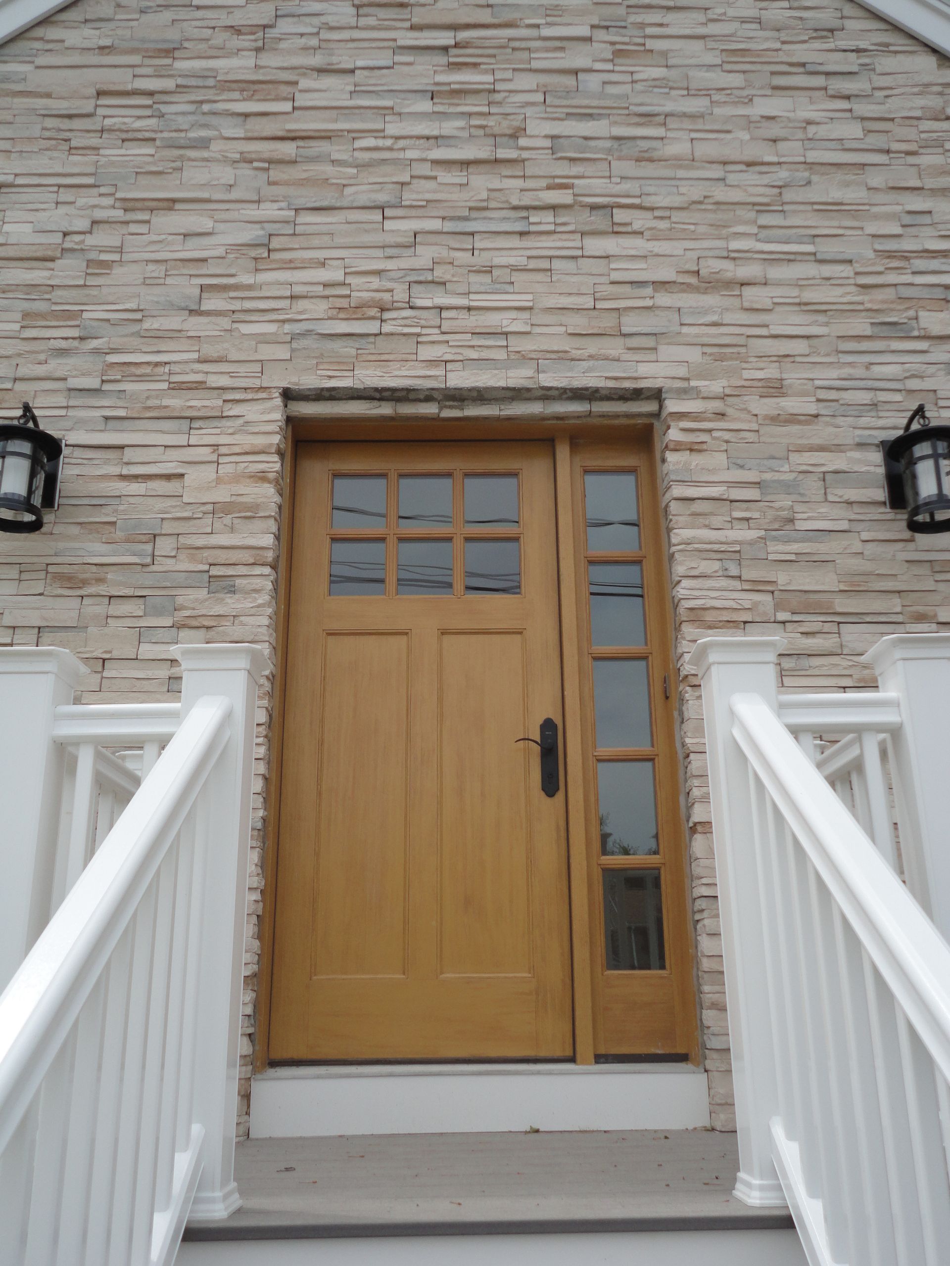 A wooden front door with a side window, flanked by white railings and black lanterns on a stone-faced exterior wall.