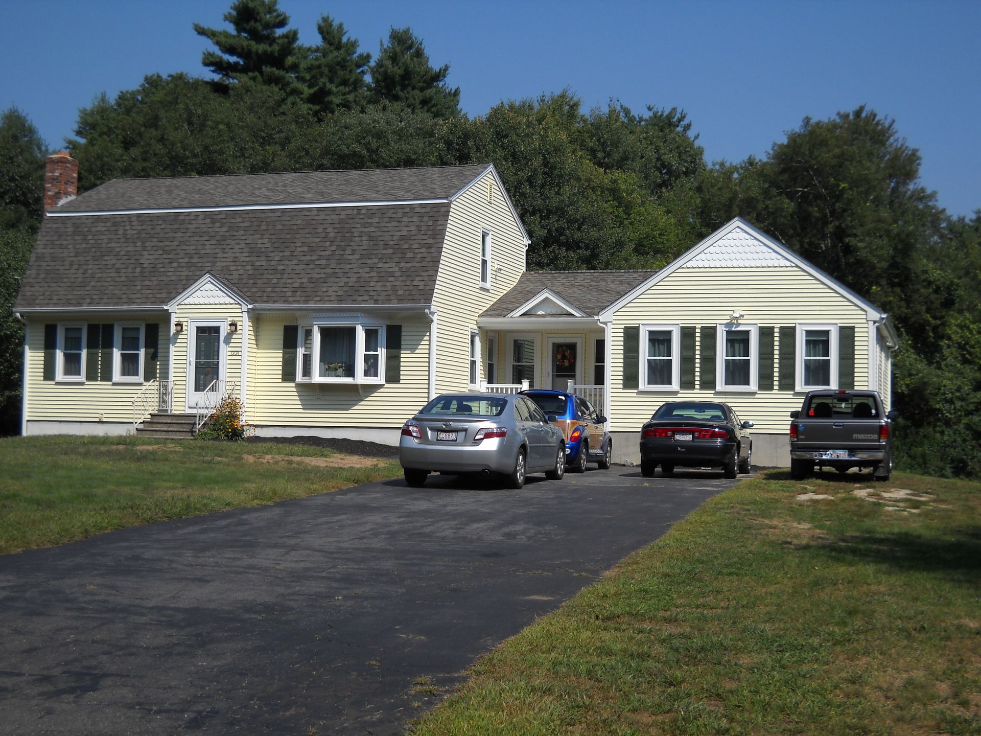 A pale yellow, two-story house with an attached garage, a paved driveway, and three parked cars on a sunny day.