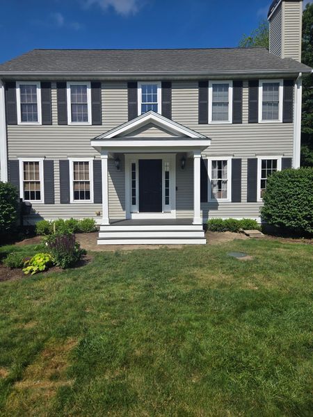 Gray colonial house with black shutters, a front porch, and green lawn.