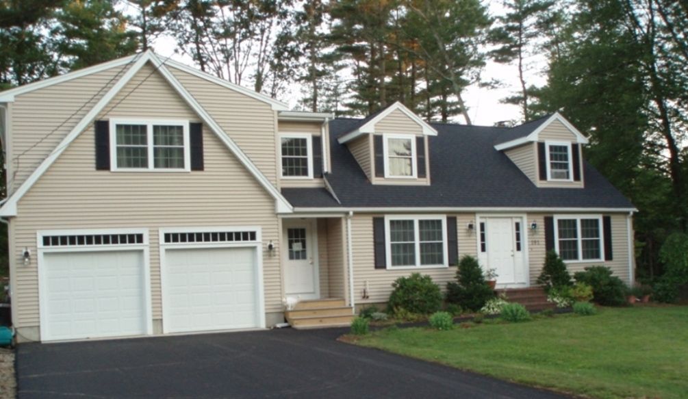 A two-story house with tan siding, a dark roof, two garage doors, and two dormer windows sits on a grassy lot.
