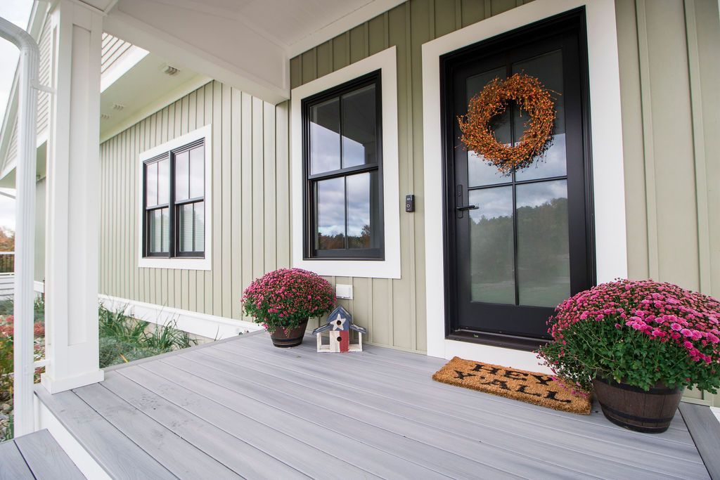 A porch with vertical sage-green siding, black-framed windows and door, two potted pink mums, and a welcome mat.