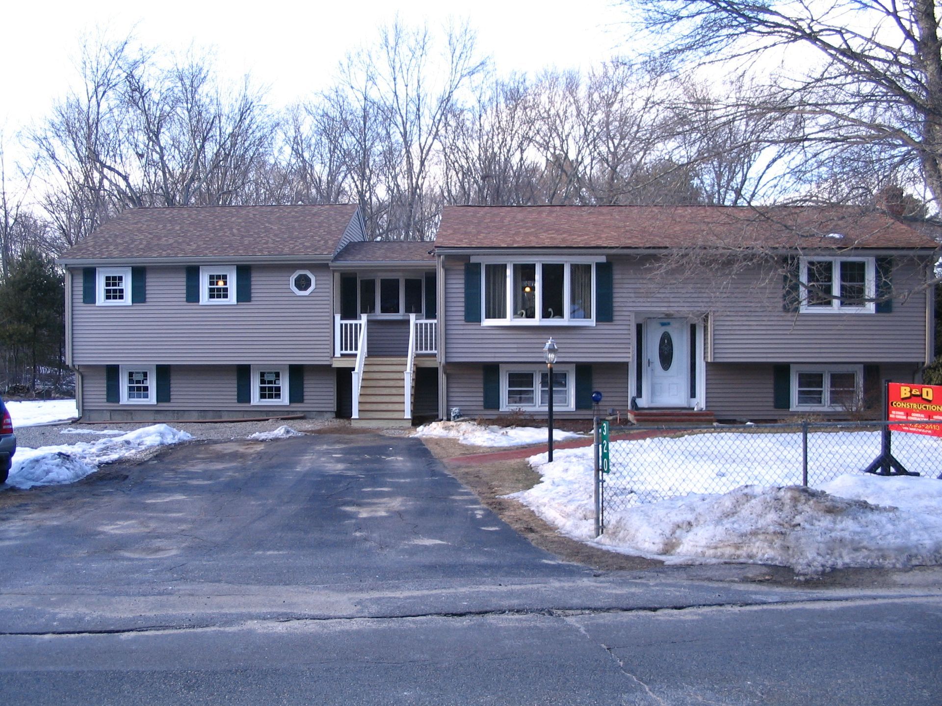 A two-story, split-level tan house with dark shutters, a front staircase, and a snowy yard with a real estate sign.