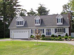 Light gray two-story house with a white garage door, three gabled dormers, and dark blue shutters on a grassy lot.