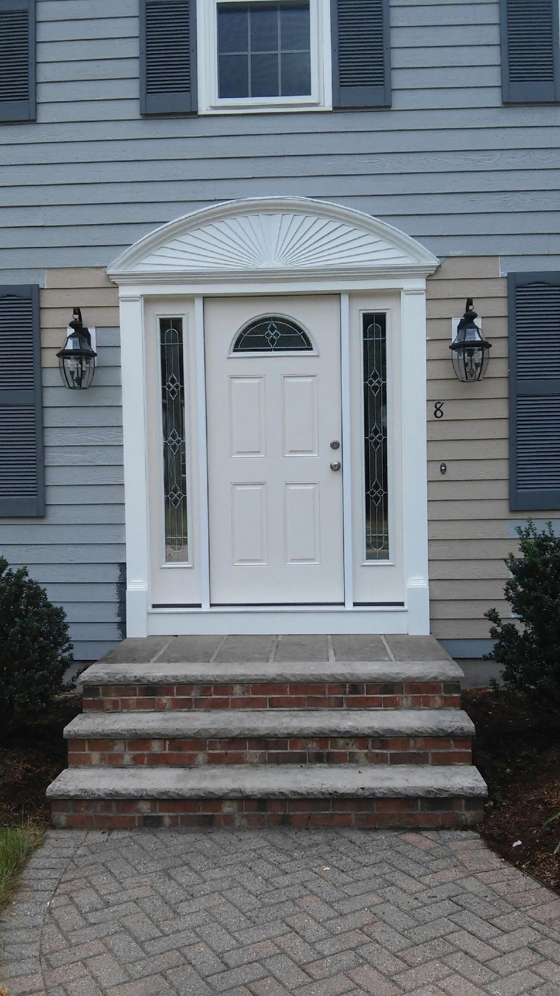 A white front door with decorative side glass panels, arched molding, and brick steps leading up from a paved walkway.