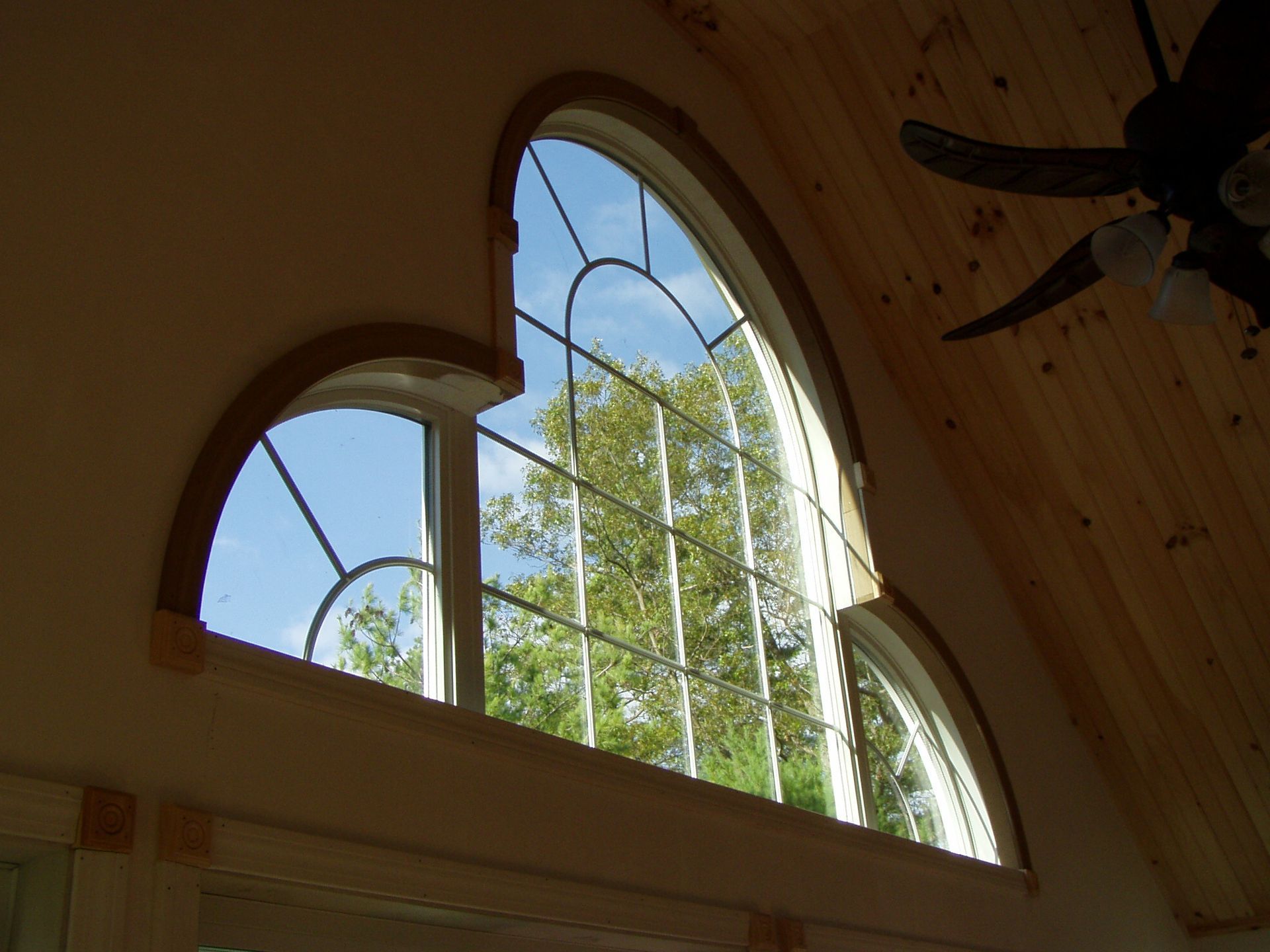 A large, multi-arched window with a grid pattern, set into a wall next to a wood-paneled ceiling with a ceiling fan.