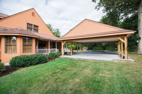 A brown house with a carport. The carport has a concrete floor, and sits next to the house. Green grass surrounds.