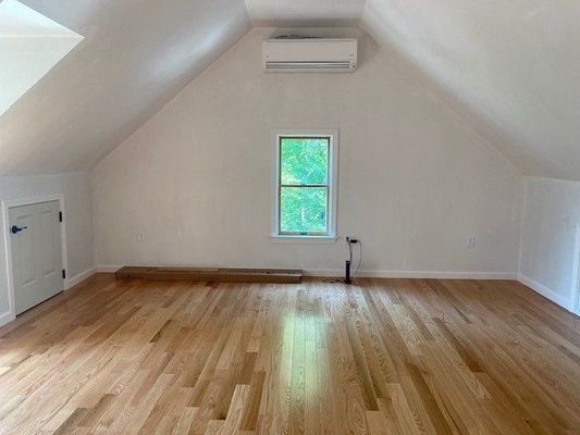 Empty attic room with hardwood floors, sloped ceilings, and a window looking out to green trees.