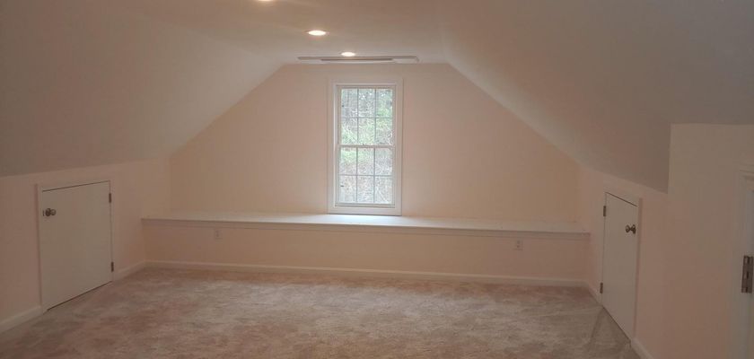 An attic room with a window, two small doors, and light-colored carpet and walls.