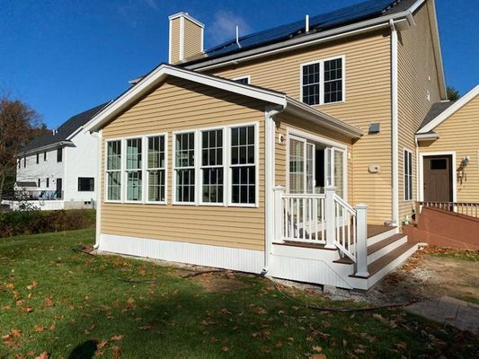 Tan house with a sunroom addition, deck, and lawn under a blue sky.