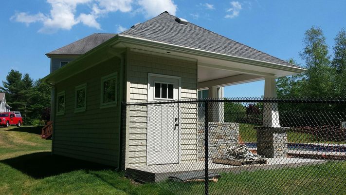 Pool house with beige siding, a gray roof, and stone columns. A chain-link fence is in the foreground.