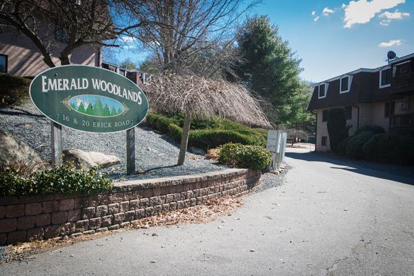 Emerald Woodlands sign on brick wall, driveway leading to buildings. Green sign with trees.