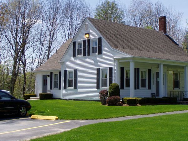White house with black shutters, green lawn, black car parked in front.