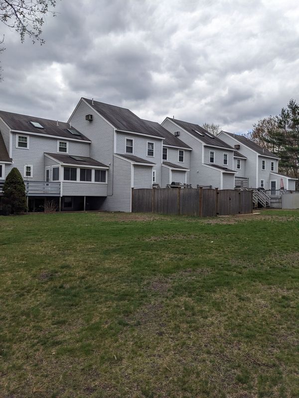 Row of gray townhouses with a fence in front, set on a grassy lawn under a cloudy sky.