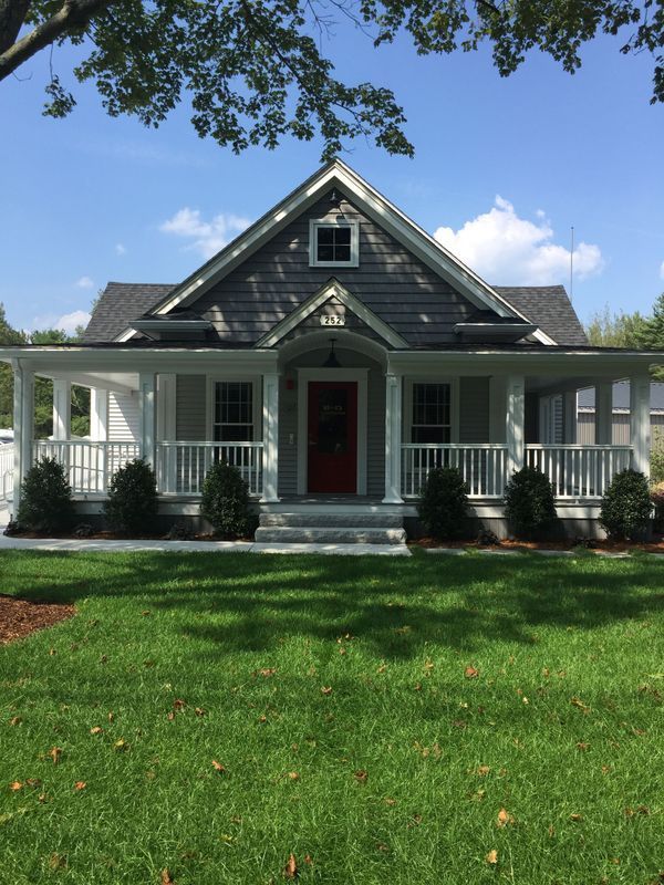 Gray and white cottage with a red door, porch, and green lawn under a blue sky.