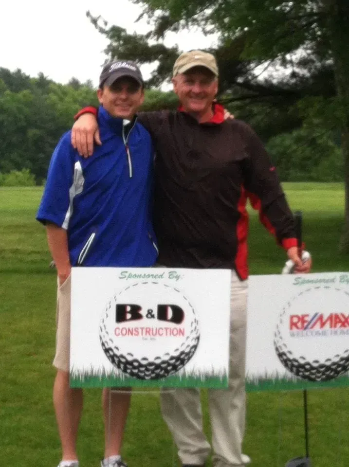 Two men at a golf outing; arm around each other. Holding signs, wearing golf attire, standing on grass.