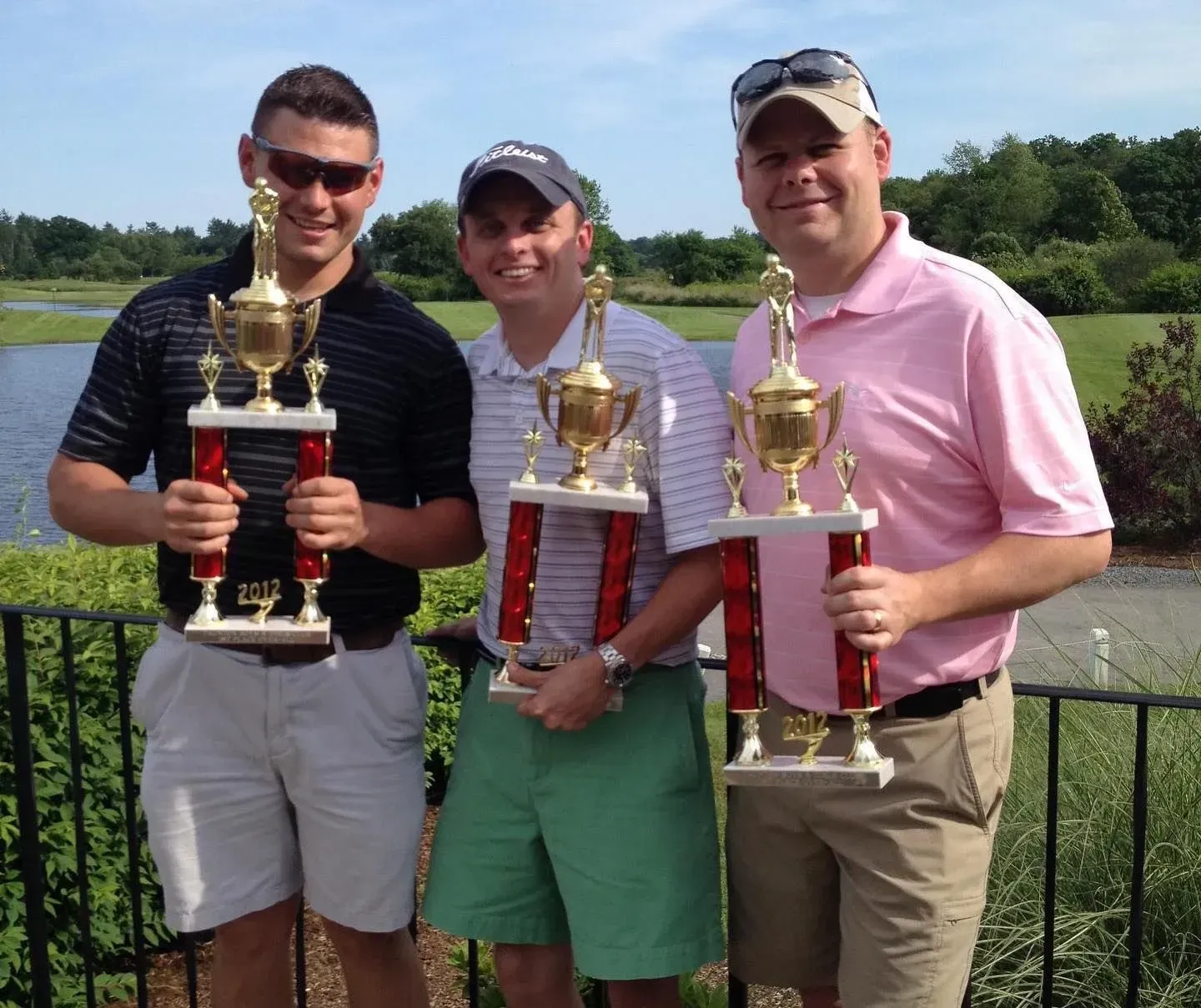 Three men holding trophies, smiling, outside near a pond.
