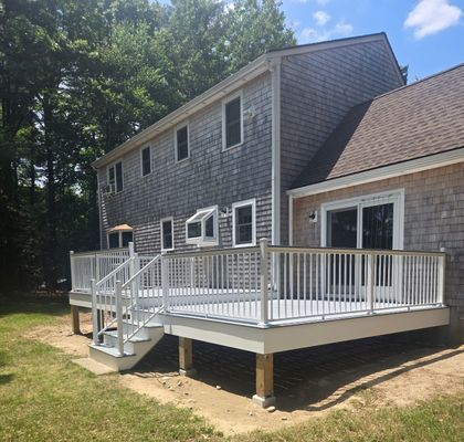 Two-story house with a gray deck and white railing. Deck steps lead to a grassy yard, sunny day.