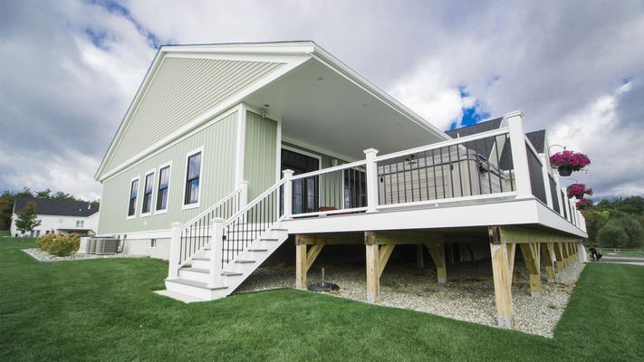 Green house with white deck, stairs, and railing. Overcast sky and green lawn.