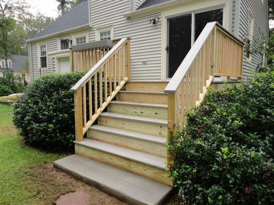 Wooden deck stairs leading to a house, with a concrete base and surrounding bushes.