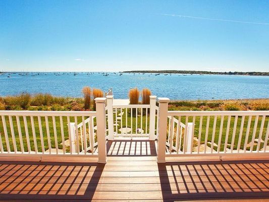 Wooden deck overlooking a blue ocean, grassy yard, and clear sky. White railing and gate.