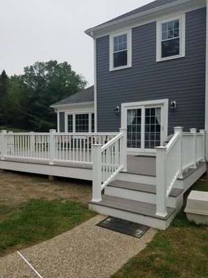 White deck with gray stairs and rails, attached to a blue-gray house with a paved walkway.