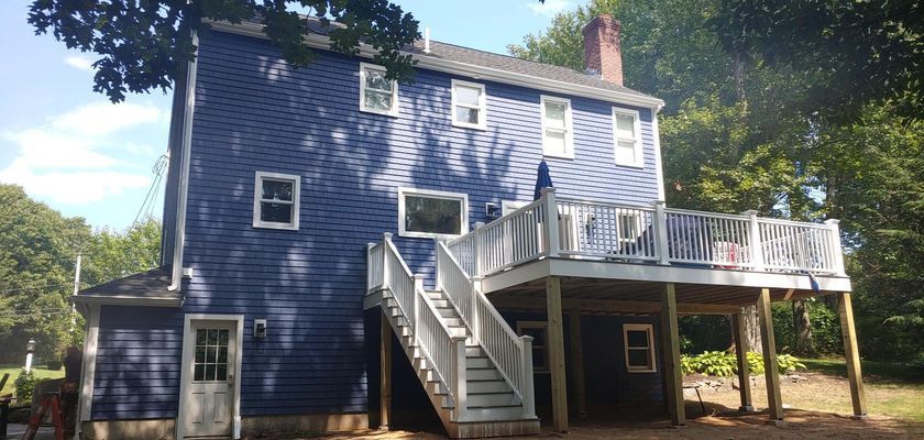 Blue house with white deck and stairs, surrounded by trees under a blue sky.