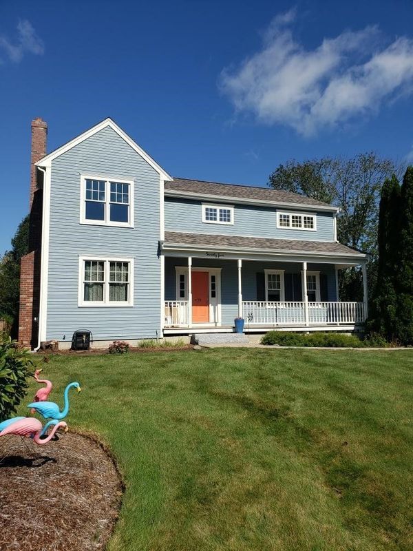 Blue house with orange door, white trim, and a wraparound porch, set on a green lawn under a blue sky.