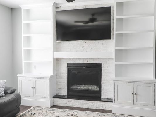Built-in white bookshelves flank a fireplace with a TV mounted above. The room is lightly colored with dark accents.