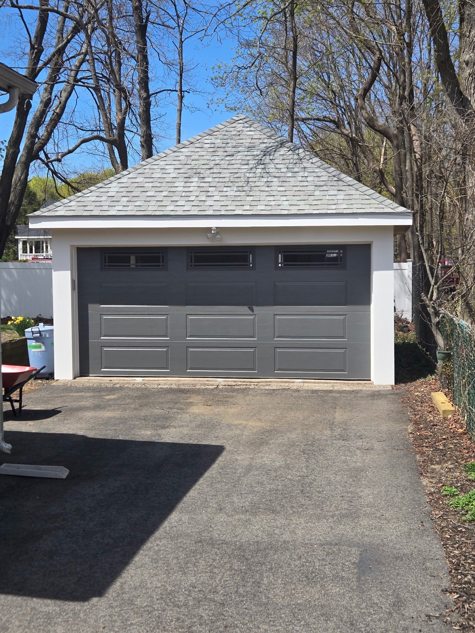 A single-car detached garage with a gray door, three windows, and a hipped roof, situated at the end of a paved driveway.