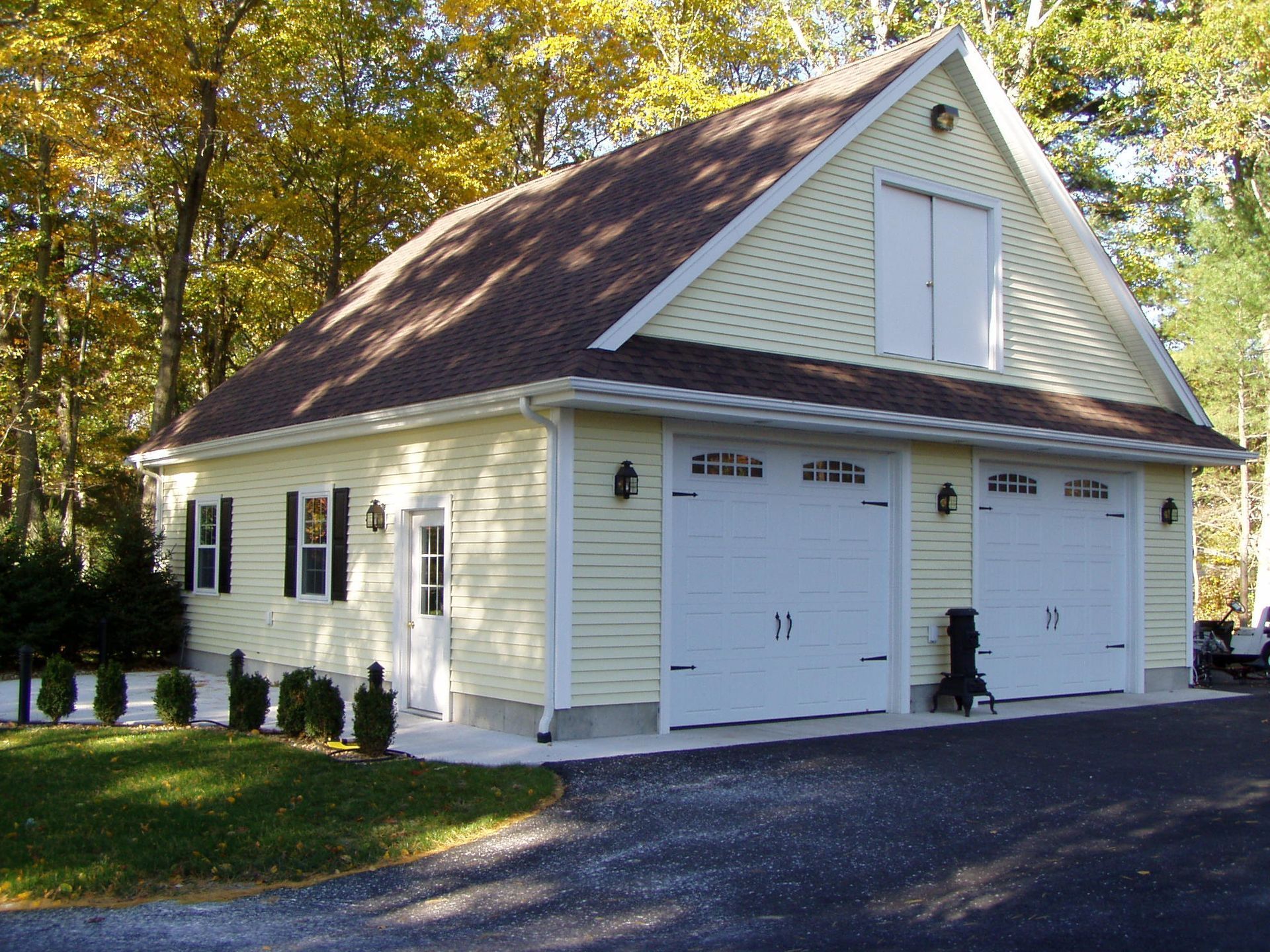 A yellow two-car garage with white trim and a loft, surrounded by autumn trees on a gravel lot.