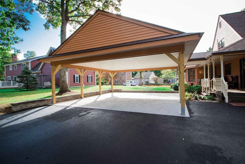 A large, open-air wooden carport with a shingled gable roof positioned over a paved driveway next to a house.