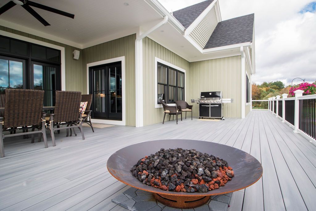 Outdoor patio with fire pit, gray deck, green house exterior, black windows, and grill.