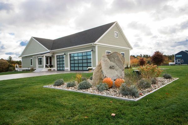 A modern, light green house with glass garage doors, large rock with pumpkins, and manicured landscaping.