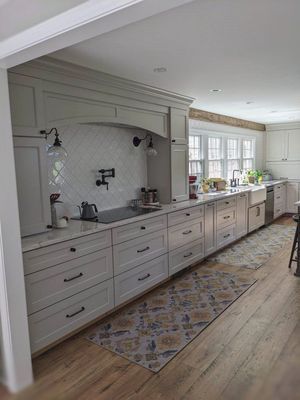 Spacious white kitchen with long cabinets, range hood, and a decorative rug on a light wood floor.