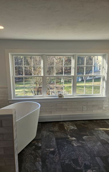 Bathroom with white tub, large windows overlooking greenery, and dark wood-look floor.