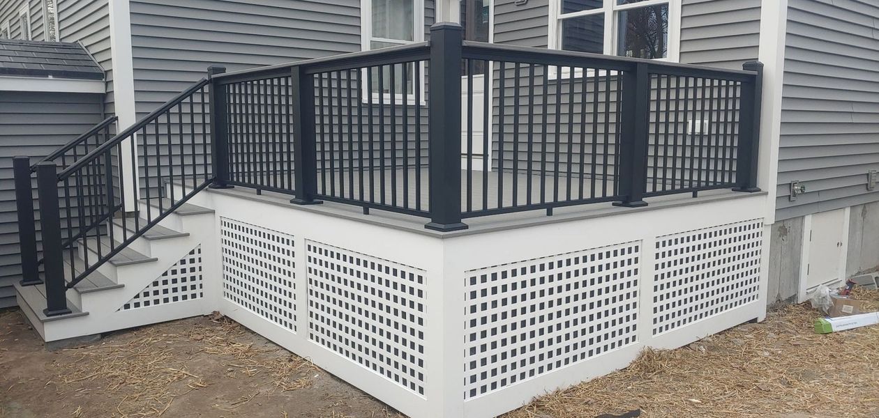 A black railing and stairs lead to a raised deck with white lattice siding, next to a gray house.