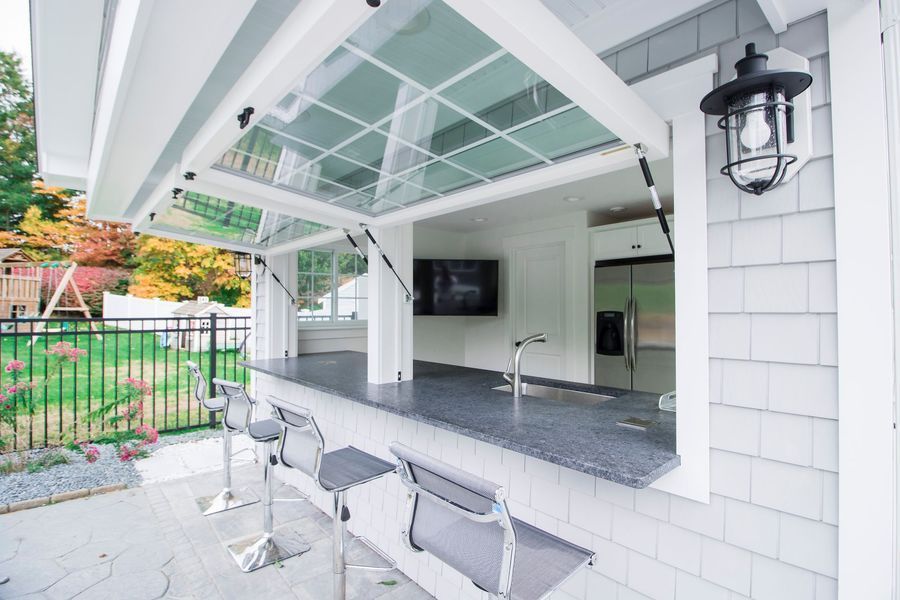 Outdoor kitchen with a folding glass window, bar seating, and a view of a yard with trees.