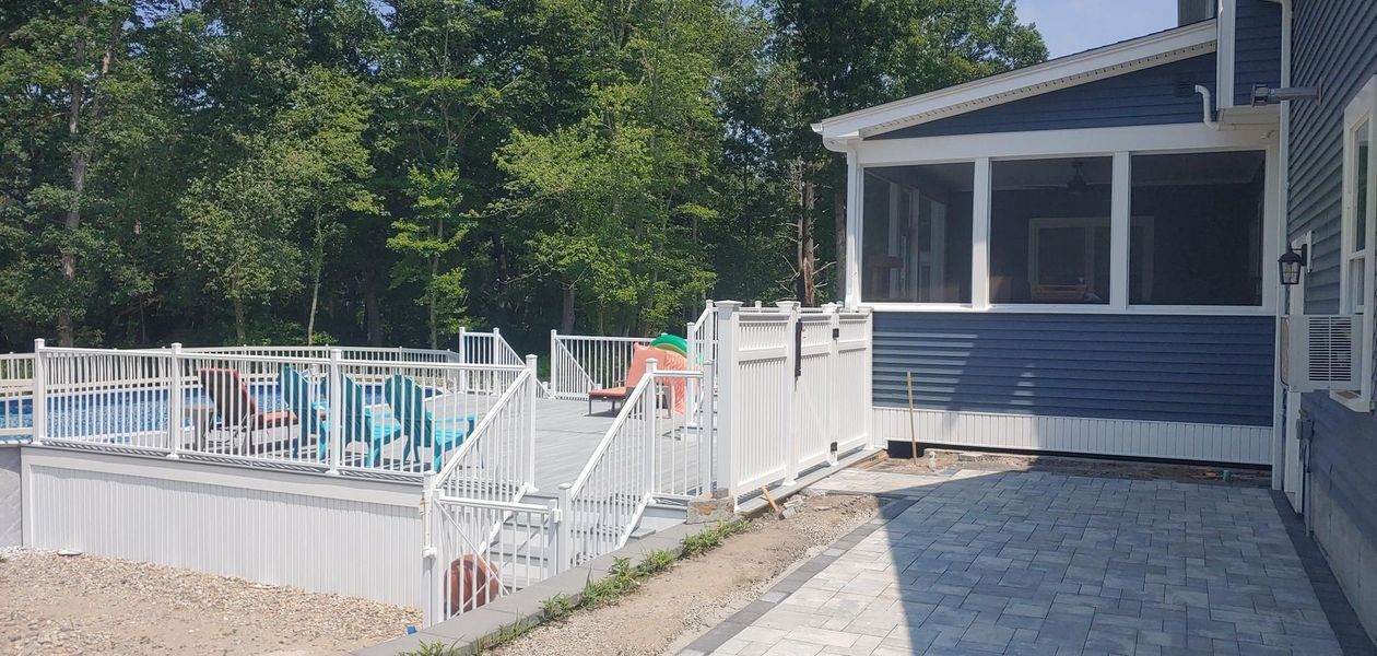 A deck with a pool and a screened porch, surrounded by trees and a paved patio.