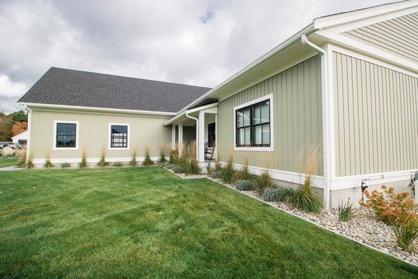 Green siding house with black windows, grass lawn, and landscaping under a cloudy sky.
