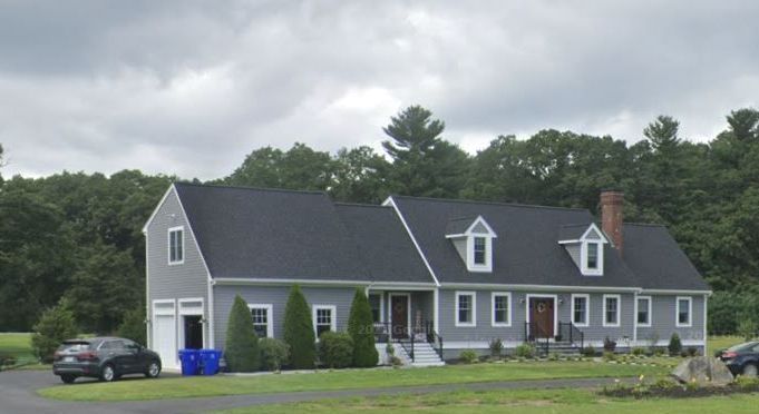 Gray house with black roof and white trim; front yard with car and trees, overcast sky.