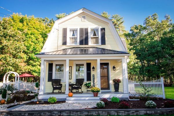 Yellow cottage with black shutters and a porch, set in a yard with white picket fence.
