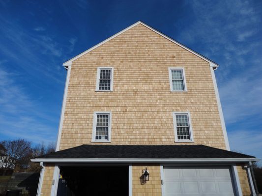 Three-story house with cedar shingle siding, white trim, and a black roof over the garage, against a blue sky.